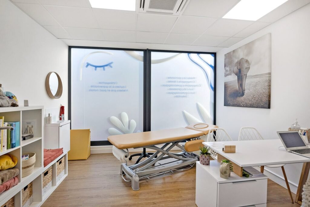 Un bureau médical ou thérapeutique moderne avec une table de traitement réglable, un bureau, des étagères avec des fournitures, et de grandes fenêtres avec des motifs en verre dépoli. Une œuvre d'art représentant un éléphant est accrochée au mur.