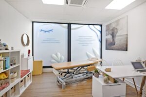 Un bureau médical ou thérapeutique moderne avec une table de traitement réglable, un bureau, des étagères avec des fournitures, et de grandes fenêtres avec des motifs en verre dépoli. Une œuvre d'art représentant un éléphant est accrochée au mur.