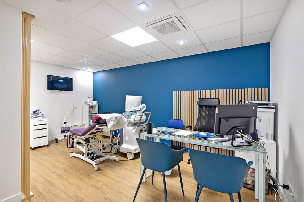 Salle d'examen médical moderne avec un fauteuil d'examen, un appareil à ultrasons, un bureau avec des chaises de bureau, un ordinateur et un mur d'accent bleu.