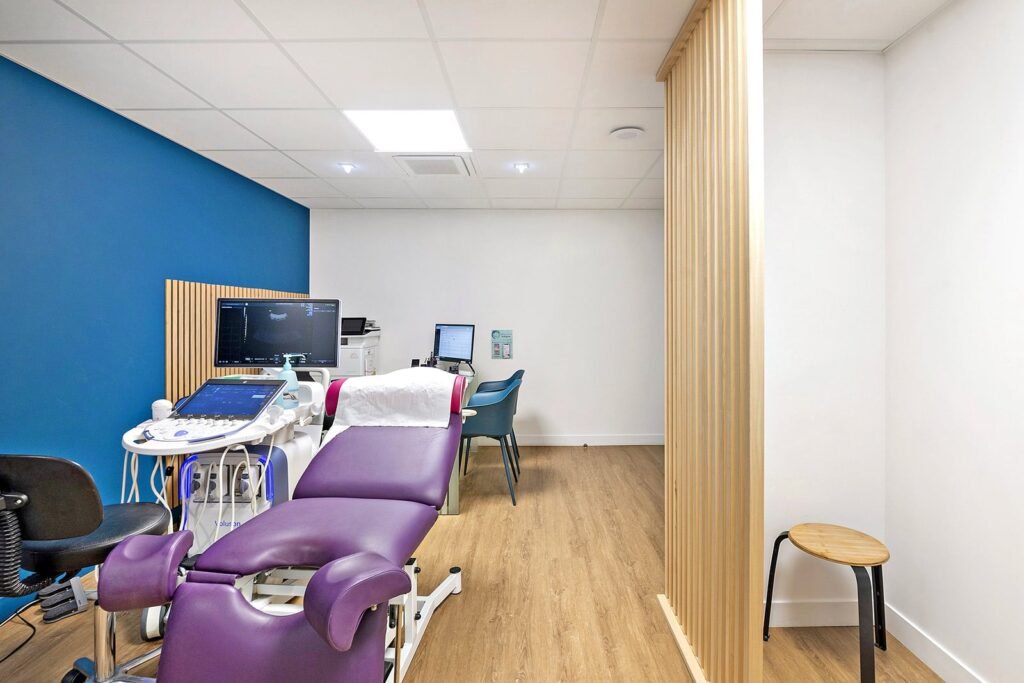 Une salle d'examen médical moderne avec une chaise d'examen violette, un appareil à ultrasons, des ordinateurs, un mur d'accent bleu, un sol en bois et une cloison de séparation en bois.