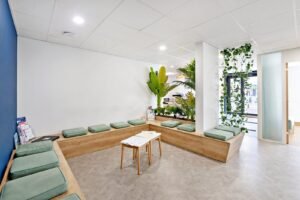 Salle d'attente moderne avec des bancs en bois, des coussins verts, des plantes d'intérieur, une petite table avec des magazines, des murs blancs et de grandes fenêtres laissant entrer la lumière naturelle.