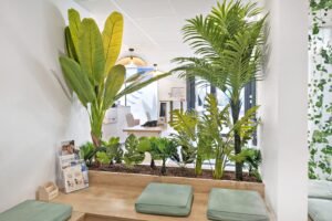 Bureau moderne avec des plantes d'intérieur, une banquette en bois avec des coussins verts et un bureau à l'arrière-plan. La lumière naturelle pénètre par de grandes fenêtres.