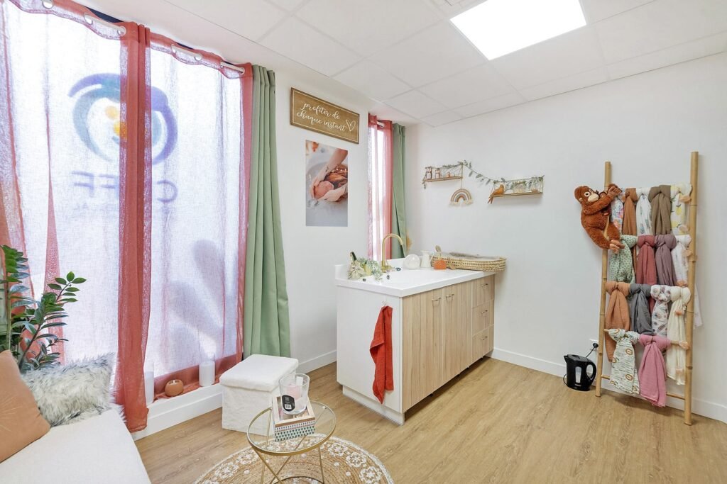 Chambre d'enfant lumineuse et organisée avec une table à langer, un tapis moelleux, une décoration murale et des langes colorés suspendus à un support en bois. La lumière naturelle filtre à travers des rideaux transparents.