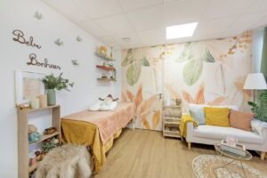 Chambre douillette avec une table de massage, un petit canapé, des feuilles, des plantes, des animaux en peluche et des étagères décoratives. Le mur porte l'inscription "Bébé Bonheur" avec des oiseaux décoratifs.