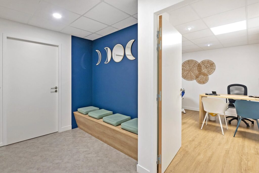 Intérieur de bureau moderne avec une petite salle d'attente dotée d'un mur d'accent bleu, de miroirs en forme de croissant de lune, d'une banquette avec des coussins et d'un bureau adjacent visible à travers une porte ouverte.
