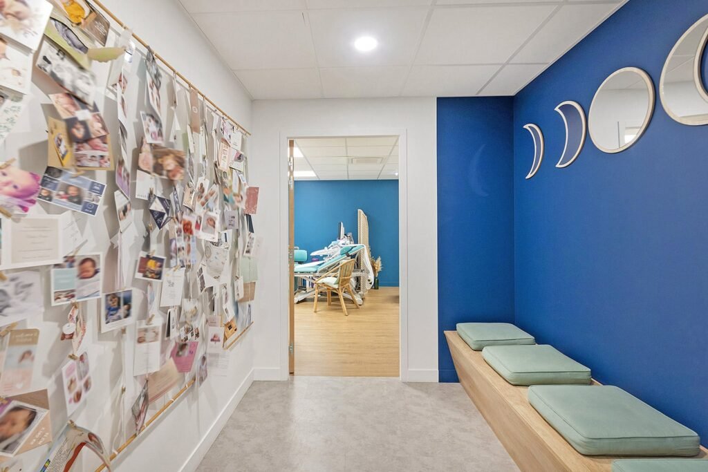 Un couloir avec un tableau d'affichage couvert de photos et de notes, un banc en bois avec des coussins verts, et une vue sur une salle avec du matériel médical ou d'examen.