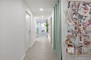 Un couloir avec des portes fermées, des lumières vives au plafond, un tableau d'affichage avec de nombreuses photos de bébés et des cartes, et une plante en pot au fond.