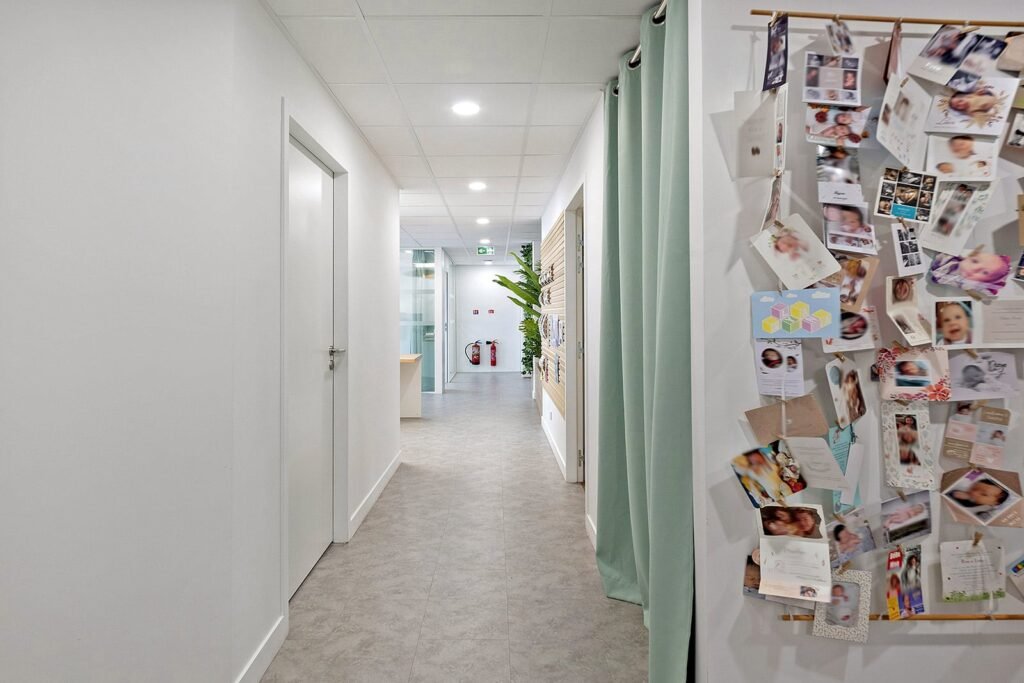 Un couloir dans un bâtiment moderne avec des murs blancs, une porte fermée à gauche et un tableau d'affichage couvert de photos et de cartes à droite.