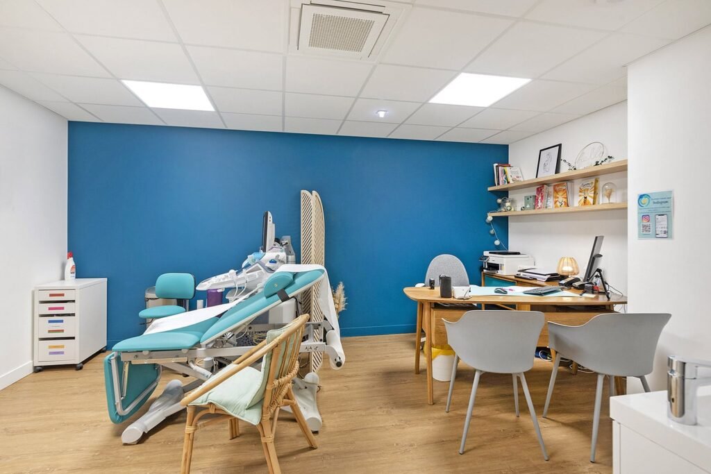 Un cabinet médical moderne avec une chaise d'examen, un bureau, un ordinateur, des étagères avec des modèles médicaux et deux chaises d'invités, le tout sur un mur d'accent bleu et un sol en bois.