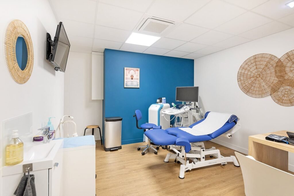 Une salle d'examen médical propre et moderne avec une chaise d'examen bleue, du matériel médical, un évier, une décoration murale et un moniteur sur un mur d'accent bleu.