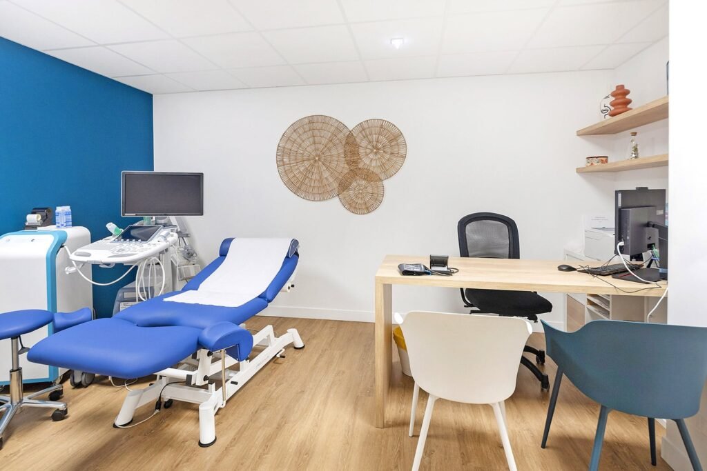 Une salle d'examen médical moderne avec une table d'examen bleue, un appareil à ultrasons, un bureau en bois avec des chaises et une décoration murale sur des murs blancs.