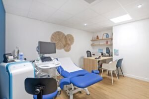 Une salle d'examen médical moderne avec une chaise d'examen bleue, un équipement d'échographie, un bureau avec des chaises et des éléments décoratifs au mur.
