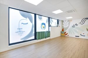 Une pièce vide et lumineuse, dotée de grandes fenêtres, où l'on trouve des dessins de visages abstraits, des tapis de yoga enroulés, des couvertures empilées et une peinture murale avec des motifs inspirés de la nature sur le mur du fond.