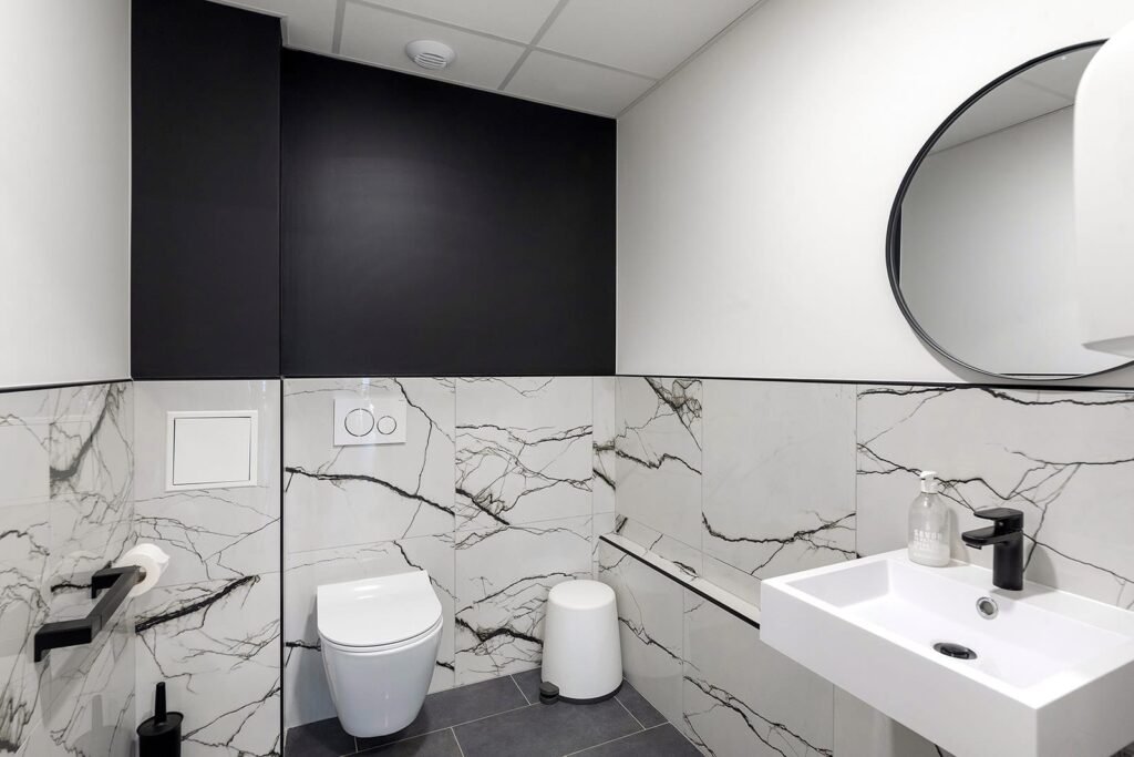 Salle de bains moderne avec toilettes murales, lavabo carré, miroir rond, carrelage marbré et luminaires noirs.