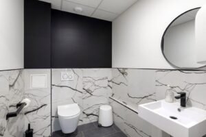 Salle de bains moderne avec toilettes murales, lavabo carré, miroir rond, carrelage marbré et luminaires noirs.
