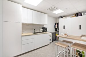 Kitchenette de bureau moderne avec des armoires blanches, des comptoirs en bois, un évier, une cafetière et une bouilloire ; table à hauteur de bar avec tabourets et placard de rangement à l'arrière-plan.