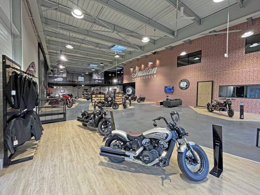 Salle d'exposition de motos spacieuse avec plusieurs motos en exposition, un mur d'accent en briques avec le logo Indian Motorcycle et des racks d'équipement de moto sur le côté.
