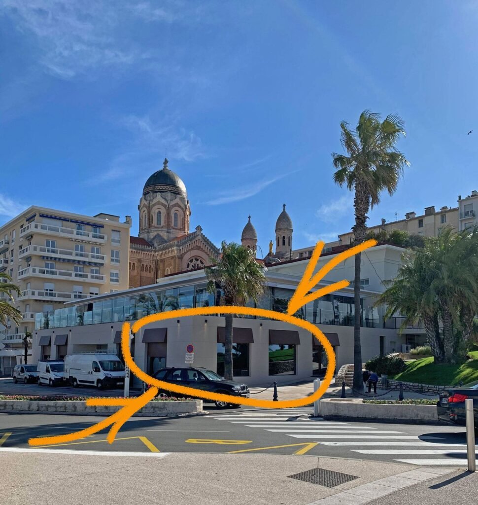 Un bâtiment en bord de mer avec de grandes fenêtres et des tours d'église en forme de dôme à l'arrière-plan ; un cercle orange et une flèche indiquent le rez-de-chaussée et la zone d'entrée.