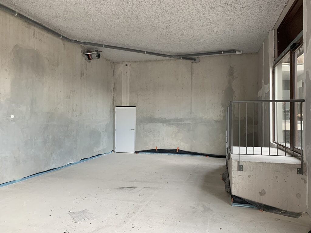 Une pièce vide et inachevée avec des murs en béton nu, une petite porte blanche, des tuyaux apparents au plafond et une rampe métallique à côté d'une zone surélevée près de grandes fenêtres.