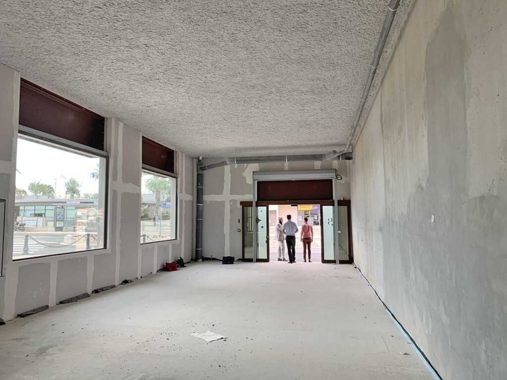 Un espace commercial vide et inachevé, aux murs nus et aux grandes fenêtres ; trois personnes se tiennent près des portes d'entrée ouvertes, tournées vers l'extérieur.