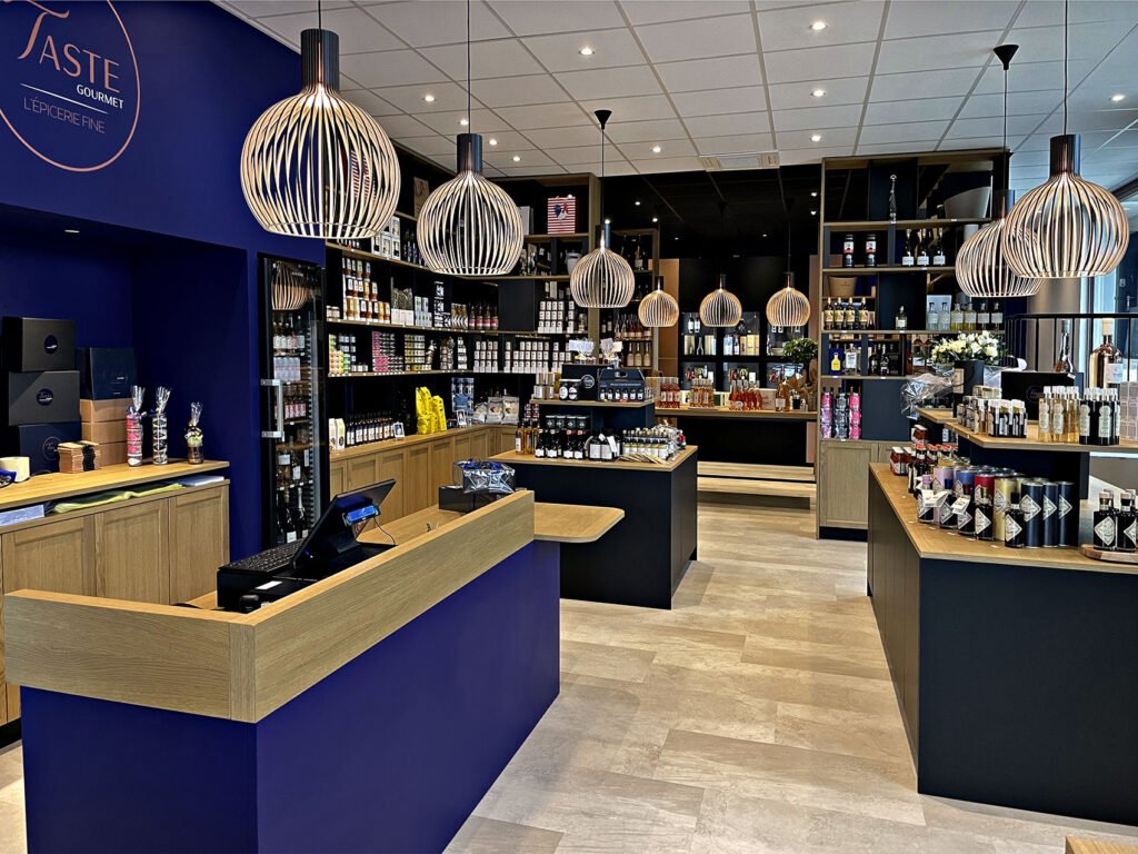 Intérieur d'une épicerie fine moderne avec des étagères en bois, des lampes suspendues et des présentoirs de produits emballés et de boissons. Un comptoir avec une caisse enregistreuse se trouve au premier plan.