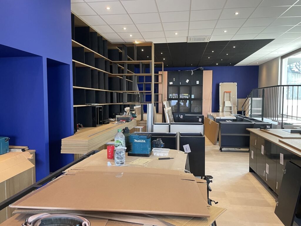 Pièce en cours de rénovation avec des cartons, des outils et des étagères ; murs bleus et noirs, cartons et matériaux de construction éparpillés dans l'espace.