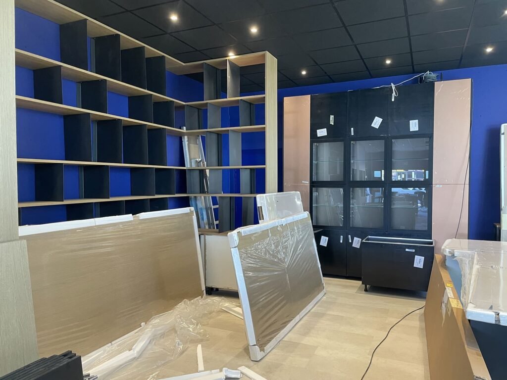Pièce avec des étagères partiellement montées, une grande armoire et des pièces de mobilier en boîte sur le sol ; murs bleus et plafond noir avec éclairage encastré.