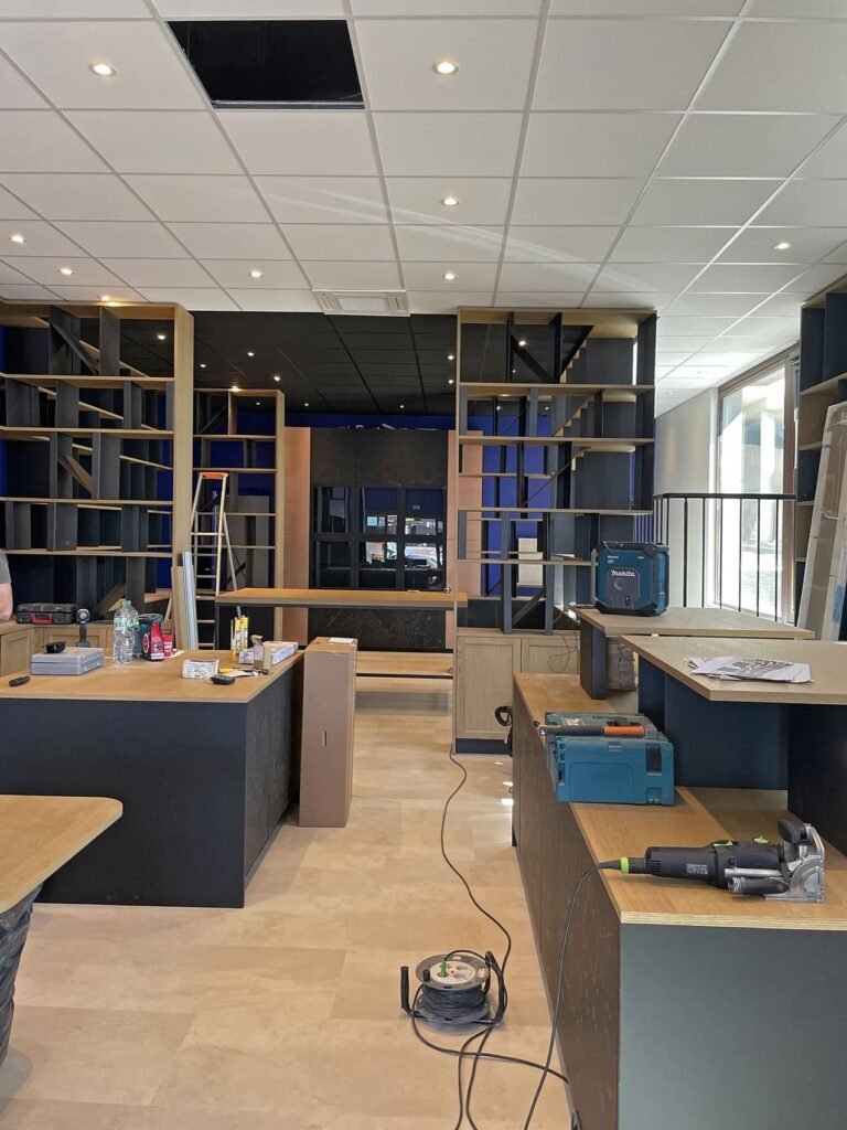 Un intérieur moderne en construction avec des étagères, des établis, des outils, une échelle et des dalles de plafond, montrant des travaux d'installation en cours.