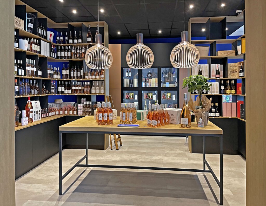 Intérieur d'un magasin de vin moderne avec des étagères de bouteilles, trois lampes suspendues et une table centrale présentant des bouteilles de vin assorties et des articles cadeaux.