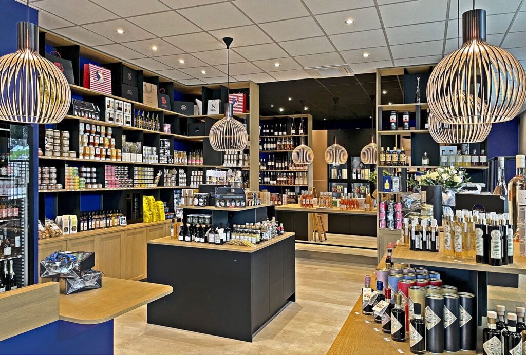 Intérieur moderne d'un magasin de vins et de produits alimentaires spécialisés, avec des étagères remplies de bouteilles, de bocaux et de produits gastronomiques, sous des lampes suspendues et un étalage bien organisé.