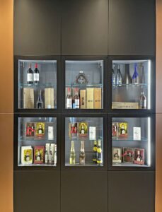 Un présentoir de bouteilles de vin et de liqueur, de coffrets cadeaux et de verrerie, disposés sur six étagères éclairées dans un meuble sombre.