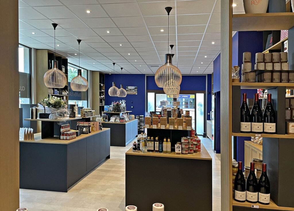 Vue intérieure d'un magasin d'alimentation spécialisée bien éclairé, avec des comptoirs gris foncé à dessus en bois, des étagères remplies de bouteilles et de bocaux, et des lampes suspendues décoratives.