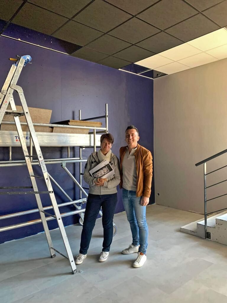 Deux personnes se tiennent dans une pièce partiellement finie avec des matériaux de construction, une échelle, un échafaudage et des escaliers visibles à l'arrière-plan.