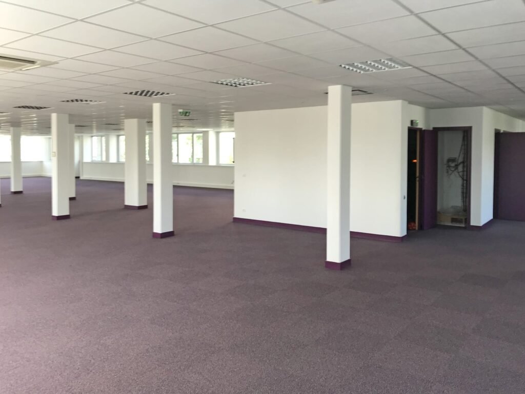 Grand espace de bureau vide aux murs blancs, à la moquette violette, aux multiples piliers et à l'éclairage fluorescent. Plusieurs portes et fenêtres sont visibles le long du mur du fond.