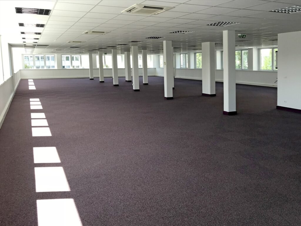 Grand espace de bureau vide avec un sol recouvert de moquette, des murs blancs, des plafonniers et de multiples colonnes de soutien ; des fenêtres s'alignent sur le côté gauche pour laisser entrer la lumière naturelle.