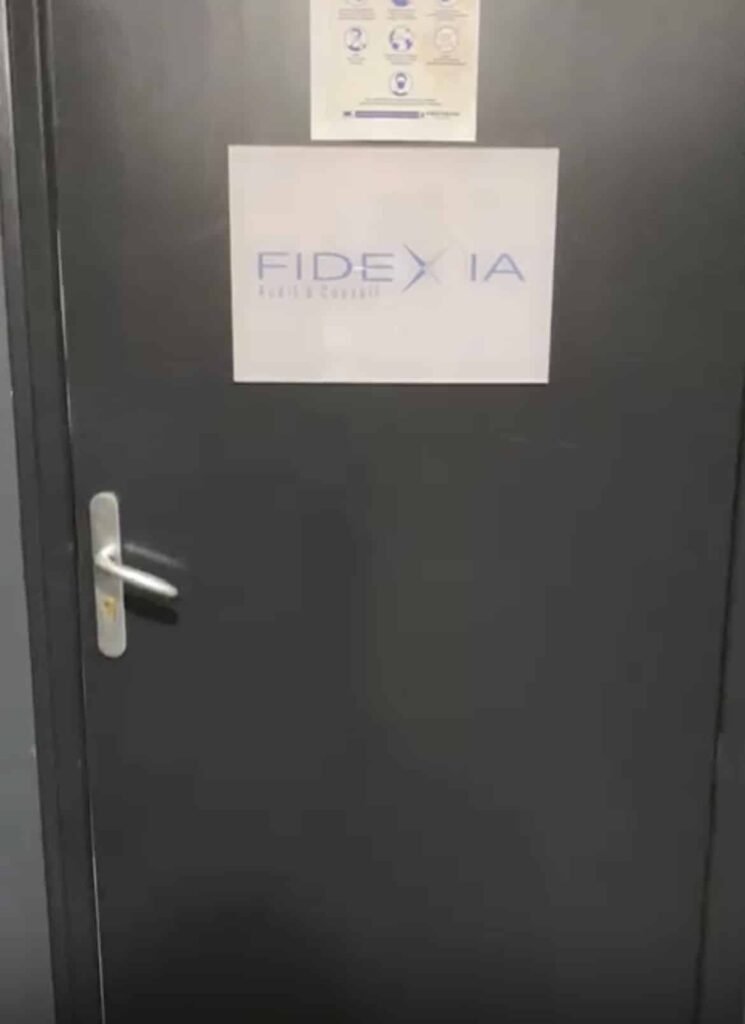 Une porte grise fermée avec une poignée argentée et un panneau portant l'inscription "FIDEXIA" en lettres bleues. Un petit panneau avec des icônes est affiché au-dessus du panneau principal.