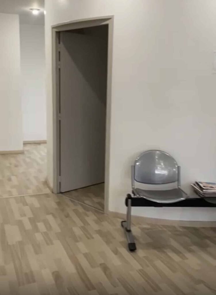 Une salle d'attente avec une chaise métallique, une pile de magazines, un sol en bois clair et une porte ouverte menant à une autre pièce.