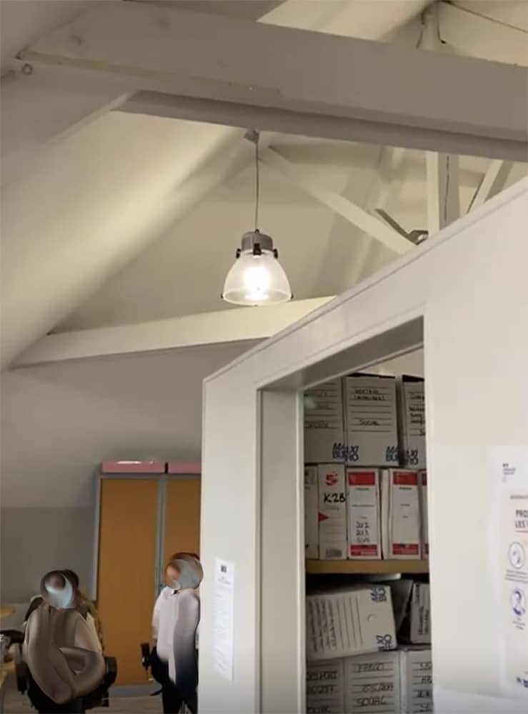 Deux personnes se tiennent dans un bureau aux murs blancs et au plafond incliné ; des étagères contenant des boîtes empilées sont visibles à l'arrière-plan.