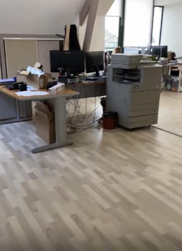 Un bureau avec des bureaux, des écrans d'ordinateur, une grande imprimante, des boîtes éparpillées et quelques personnes travaillant à l'arrière-plan.