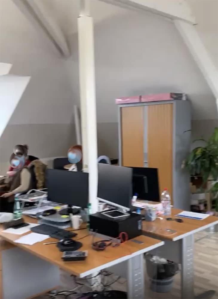Espace de bureau moderne avec des bureaux, des ordinateurs et des fournitures de bureau ; quelques personnes travaillent à leur bureau ; de grandes armoires et une plante sont visibles à l'arrière-plan.