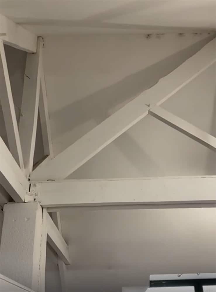 Des poutres et des fermes apparentes peintes en blanc se croisent en angle avec un plafond et un mur blancs.