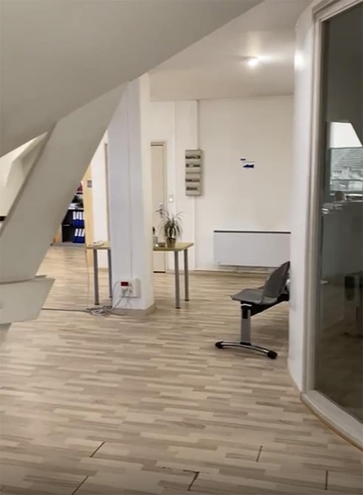 Bureau moderne avec un sol en bois clair, une petite table avec une plante, une rangée de chaises et une pièce vitrée sur la droite. Les murs et le décor sont minimalistes.