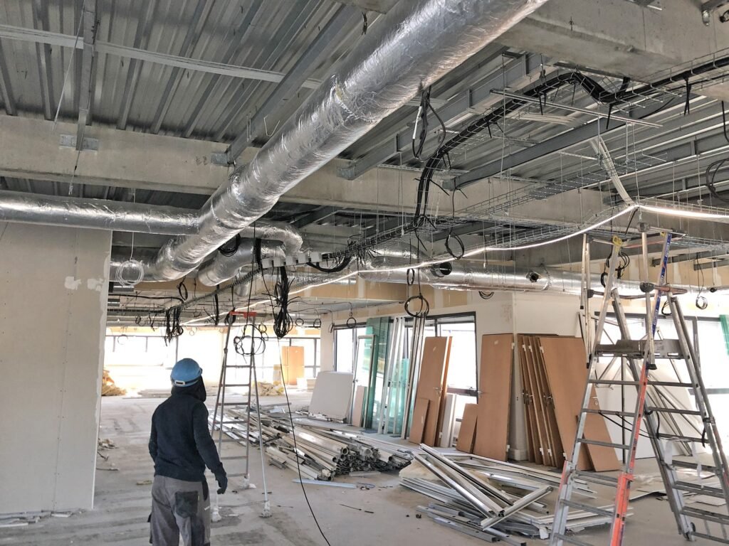 Un ouvrier de la construction se tient dans un bâtiment inachevé où sont exposés des conduits, des câbles électriques, des échelles et des matériaux de construction éparpillés sur le sol.