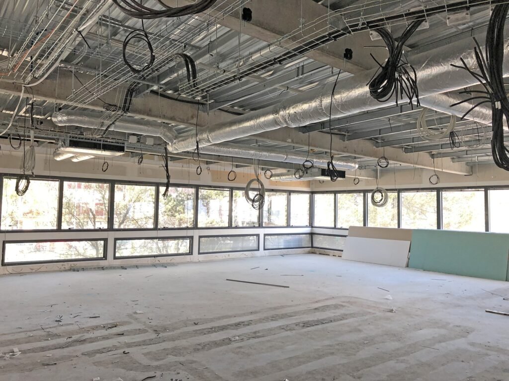 Espace de bureau inachevé avec des conduits apparents, des fils suspendus, de grandes fenêtres et des matériaux de construction sur le sol.