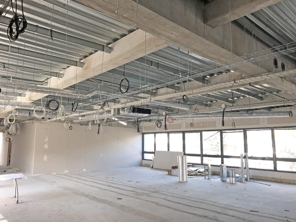 Intérieur d'un bâtiment en construction avec des poutres de plafond apparentes, des fils suspendus, des conduits, des murs et un sol non finis. De grandes fenêtres bordent un côté de la pièce.