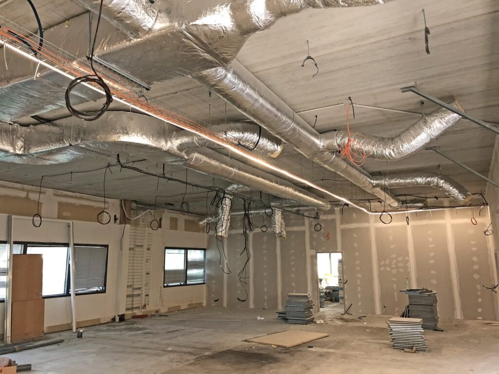 Intérieur d'un bâtiment en construction avec des conduits exposés, des câbles, des murs non finis et des piles de tuiles sur le sol.