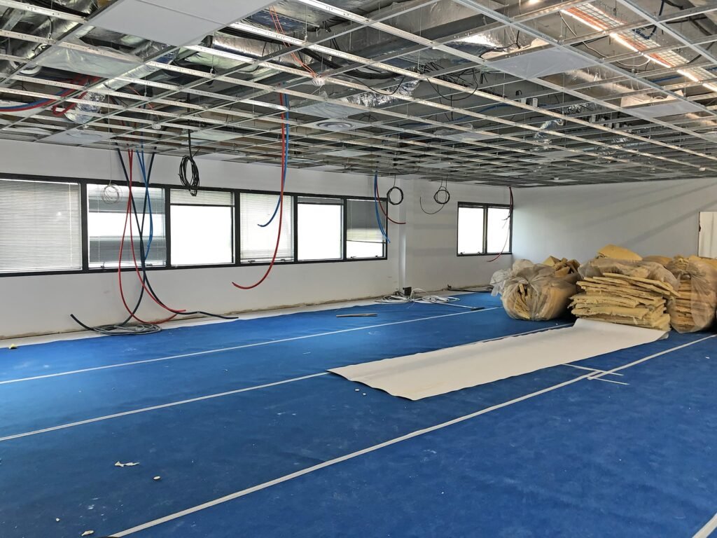 Espace de bureaux en construction avec un plafond apparent, des câbles suspendus, un revêtement de sol bleu, des fenêtres et des piles de matériaux d'isolation sur le côté droit.