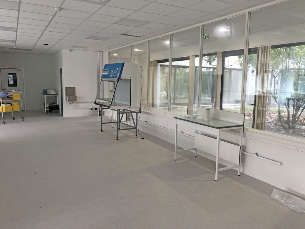 Une salle de laboratoire pratiquement vide avec une armoire de sécurité biologique, des tables et de grandes fenêtres donnant sur les arbres d'une cour extérieure.