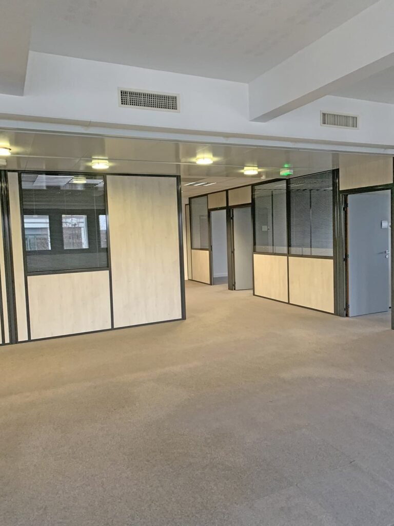 Espace de bureau vide avec moquette beige, cloisons en bois clair et en verre, et plafonniers. Plusieurs portes et fenêtres sont visibles le long des pièces cloisonnées.