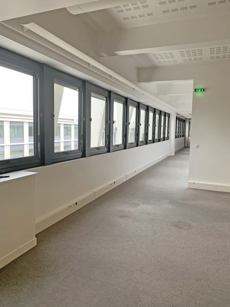 Un long couloir vide avec une moquette grise, de grandes fenêtres d'un côté, des murs blancs et un panneau de sortie vert à l'extrémité.